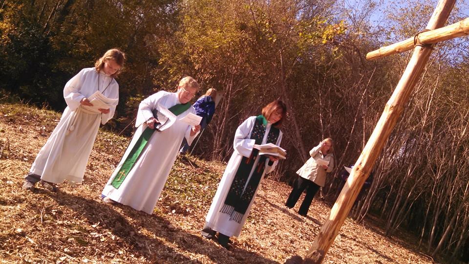 Priest, and others in liturgical robes standing at cross outside.